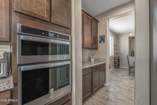 kitchen featuring double oven, wood tiled floor, dark brown cabinetry, and light stone counters