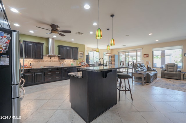 kitchen featuring a center island with sink, a kitchen breakfast bar, stainless steel refrigerator, wall chimney exhaust hood, and decorative light fixtures