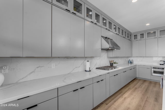 kitchen with light stone counters, light hardwood / wood-style floors, range hood, decorative backsplash, and stainless steel appliances