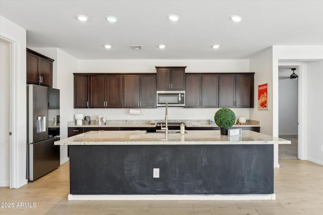 kitchen with a kitchen island with sink, dark brown cabinetry, stainless steel appliances, visible vents, and light stone countertops