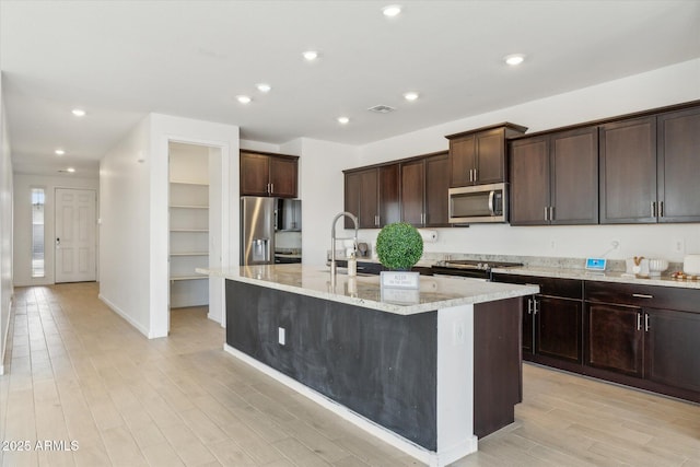 kitchen with stainless steel appliances, light wood finished floors, an island with sink, and dark brown cabinetry