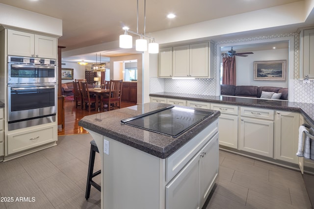 kitchen featuring double oven, black electric stovetop, a ceiling fan, backsplash, and a center island