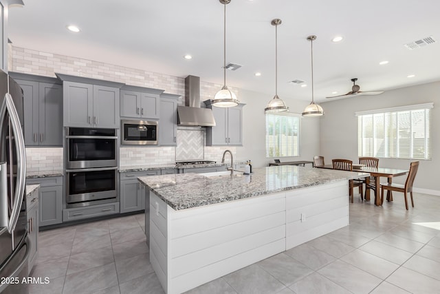 kitchen with sink, hanging light fixtures, appliances with stainless steel finishes, an island with sink, and wall chimney range hood