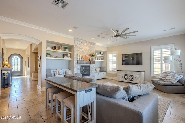 living room with a stone fireplace, crown molding, built in features, and ceiling fan