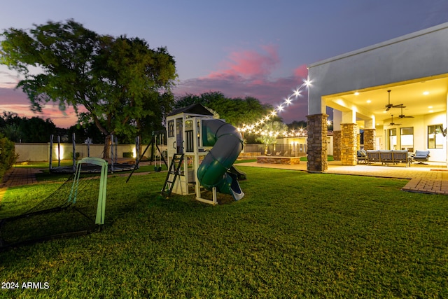 playground at dusk with a lawn, outdoor lounge area, a trampoline, ceiling fan, and a patio area