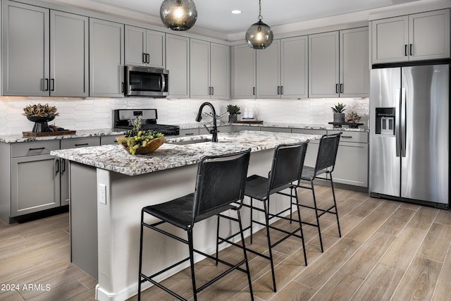 kitchen with light stone countertops, stainless steel appliances, a center island with sink, light hardwood / wood-style flooring, and hanging light fixtures