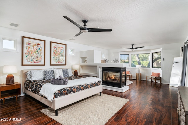 bedroom featuring a multi sided fireplace, a textured ceiling, ceiling fan, and dark wood-type flooring