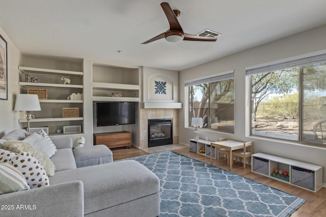 living area featuring built in shelves, visible vents, a ceiling fan, a tile fireplace, and wood finished floors