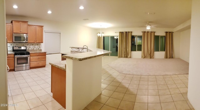 kitchen featuring an island with sink, light tile patterned flooring, backsplash, stainless steel appliances, and ceiling fan with notable chandelier