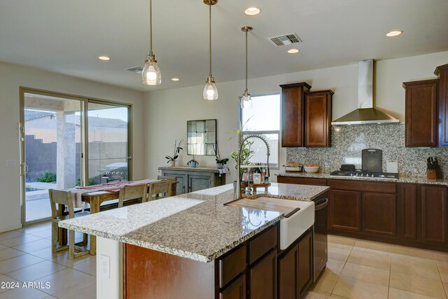 kitchen featuring light stone counters, tasteful backsplash, an island with sink, wall chimney exhaust hood, and light tile patterned floors