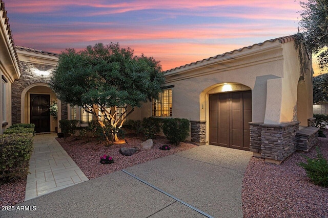 exterior entry at dusk featuring stucco siding, an attached garage, stone siding, driveway, and a tiled roof