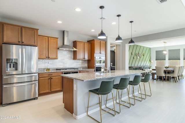 kitchen featuring a kitchen bar, an island with sink, appliances with stainless steel finishes, pendant lighting, and wall chimney exhaust hood