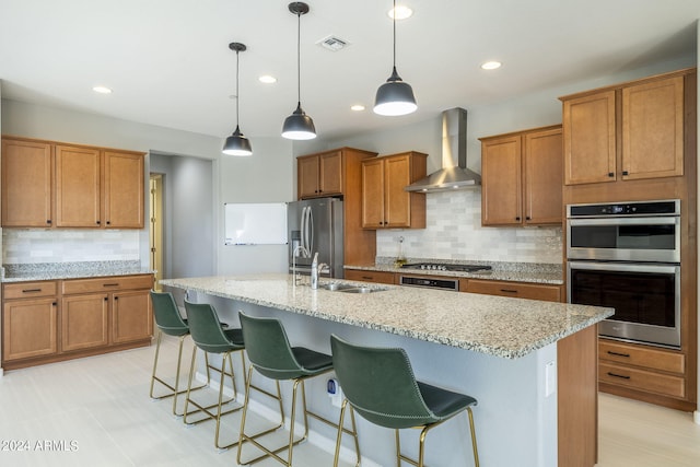 kitchen featuring decorative light fixtures, wall chimney range hood, appliances with stainless steel finishes, and a kitchen island with sink