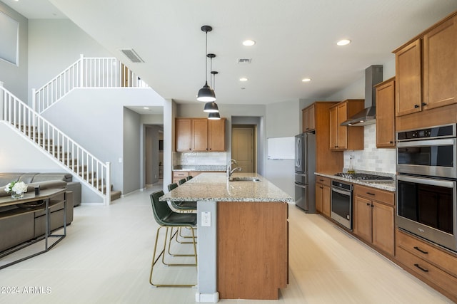 kitchen featuring light stone countertops, appliances with stainless steel finishes, wall chimney exhaust hood, an island with sink, and sink