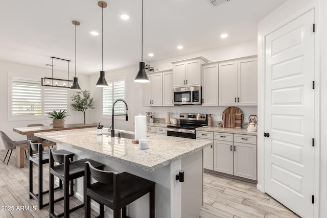 kitchen with a kitchen island with sink, decorative light fixtures, appliances with stainless steel finishes, light wood-type flooring, and light stone counters