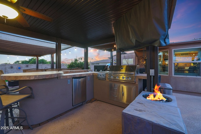 patio terrace at dusk with ceiling fan, area for grilling, an outdoor fire pit, and exterior kitchen