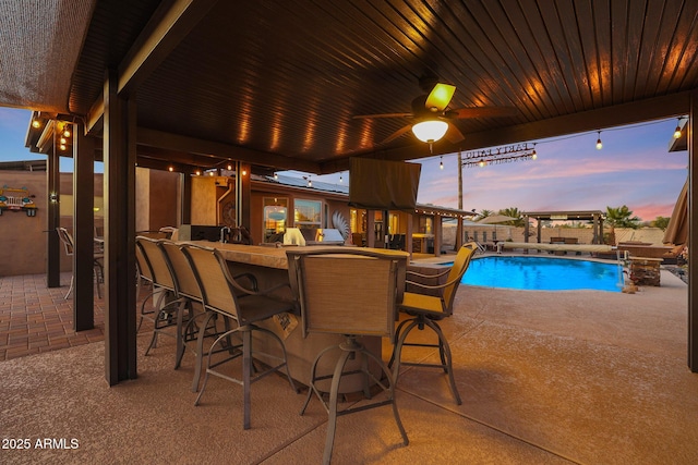 pool at dusk featuring ceiling fan, a bar, and a patio