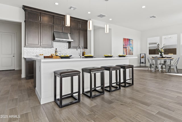 kitchen featuring a breakfast bar, pendant lighting, decorative backsplash, dark brown cabinetry, and a center island with sink