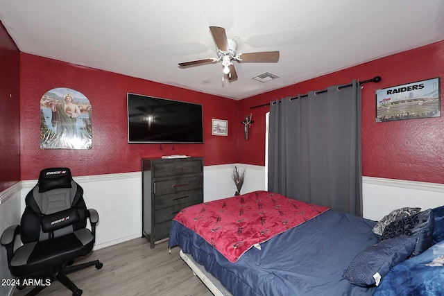 bedroom featuring a ceiling fan, wood finished floors, visible vents, a wainscoted wall, and a textured wall