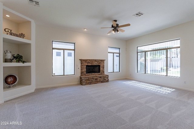 unfurnished living room with ceiling fan, built in shelves, a stone fireplace, and light colored carpet