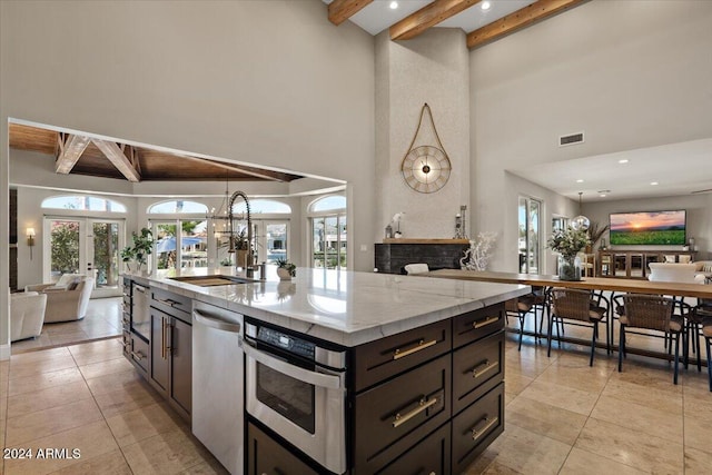 kitchen featuring beamed ceiling, an island with sink, stainless steel appliances, sink, and light stone counters