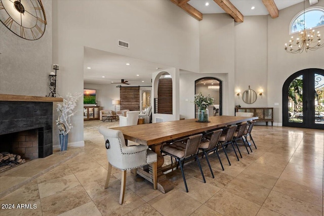 dining room with ceiling fan with notable chandelier, french doors, a tiled fireplace, a high ceiling, and beam ceiling