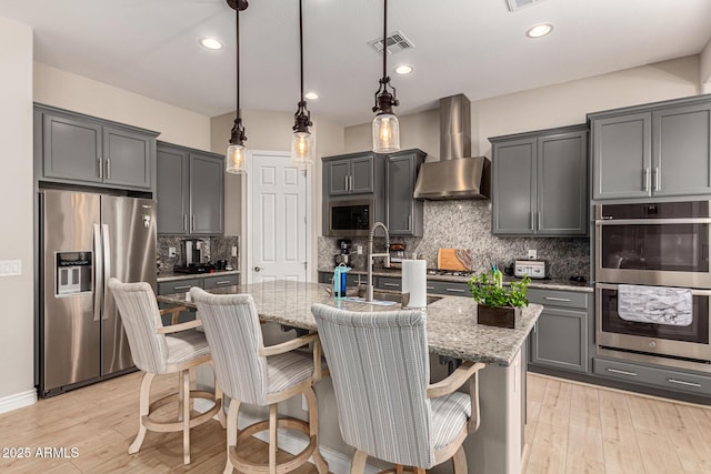 kitchen featuring appliances with stainless steel finishes, an island with sink, hanging light fixtures, light stone countertops, and wall chimney exhaust hood