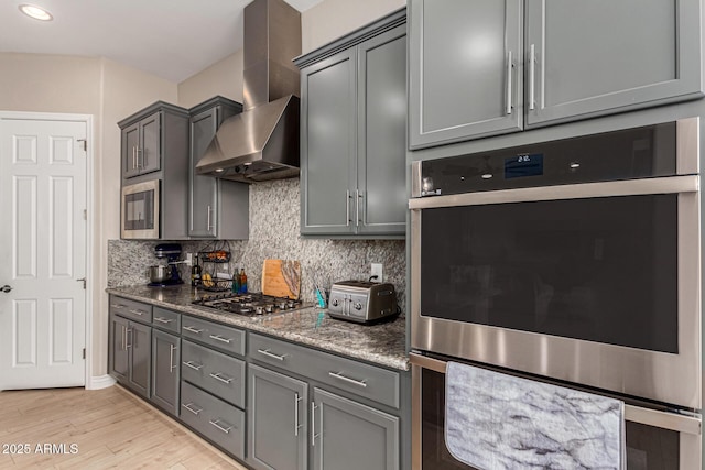 kitchen featuring gray cabinetry, wall chimney range hood, dark stone counters, and appliances with stainless steel finishes