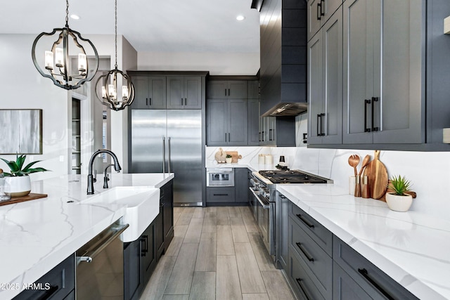 kitchen with light stone countertops, a notable chandelier, a sink, wall chimney exhaust hood, and premium appliances