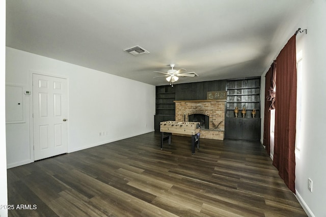 living room featuring ceiling fan, built in features, dark wood-type flooring, and a brick fireplace