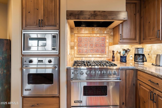 kitchen featuring light stone counters, custom range hood, appliances with stainless steel finishes, brown cabinets, and backsplash