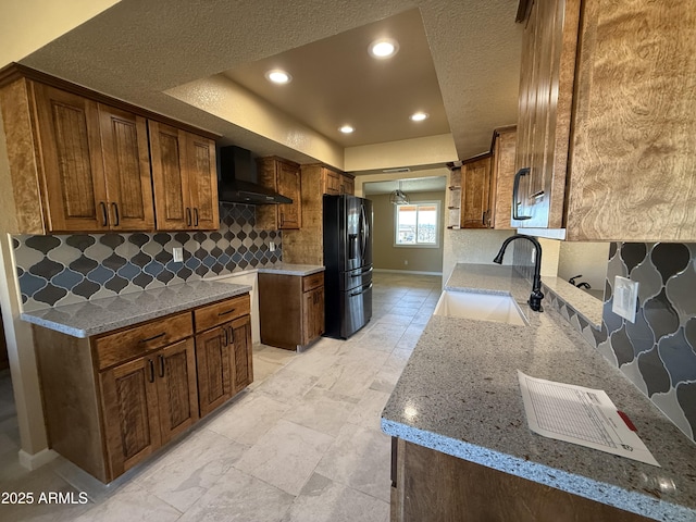 kitchen with light stone counters, a sink, black fridge with ice dispenser, a raised ceiling, and wall chimney exhaust hood
