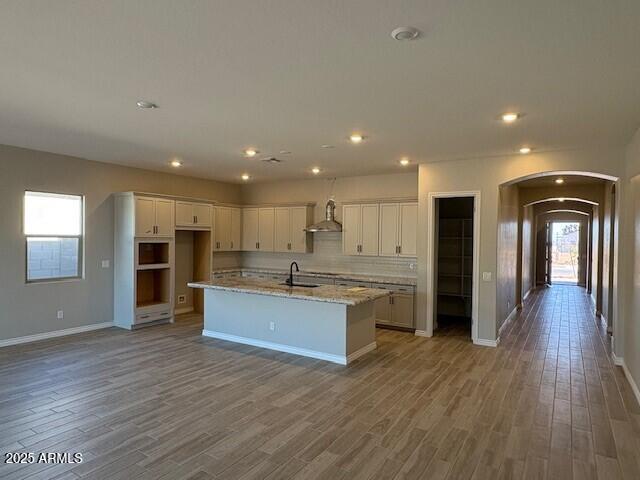 kitchen featuring arched walkways, an island with sink, a sink, white cabinetry, and wall chimney exhaust hood