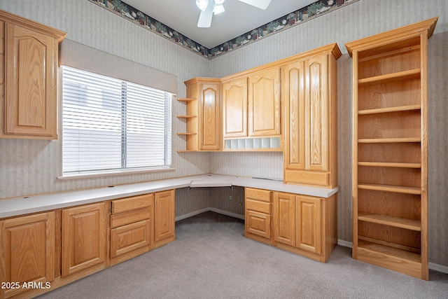 unfurnished office featuring baseboards, wallpapered walls, light carpet, built in study area, and ceiling fan