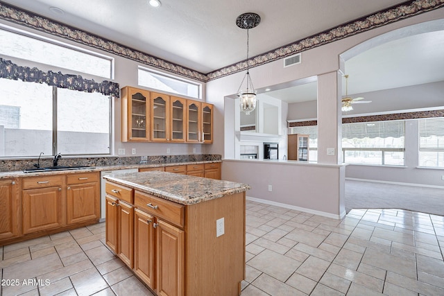 kitchen with ceiling fan with notable chandelier, glass insert cabinets, a kitchen island, light stone counters, and a sink