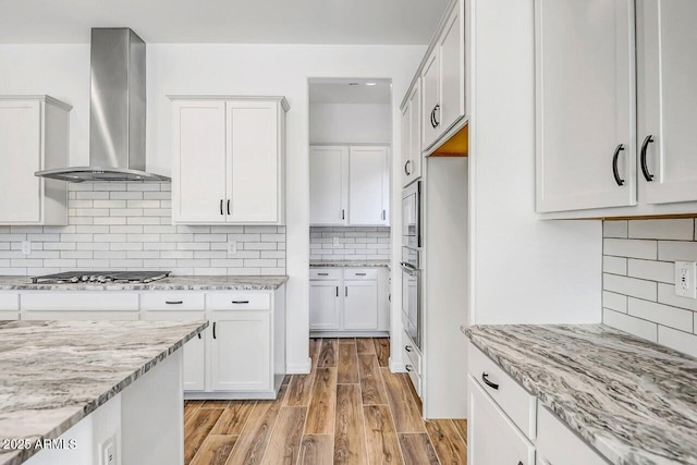 kitchen with white cabinets, stainless steel appliances, light stone countertops, light wood-type flooring, and wall chimney exhaust hood