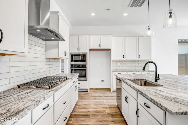 kitchen featuring appliances with stainless steel finishes, sink, white cabinets, hanging light fixtures, and wall chimney range hood