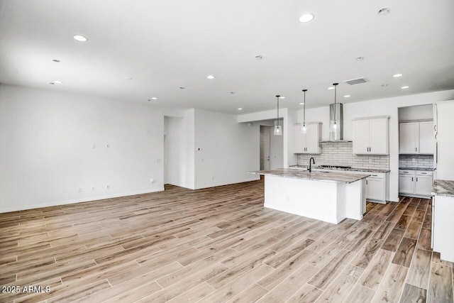 kitchen with a kitchen island with sink, light stone countertops, white cabinets, decorative light fixtures, and wall chimney exhaust hood