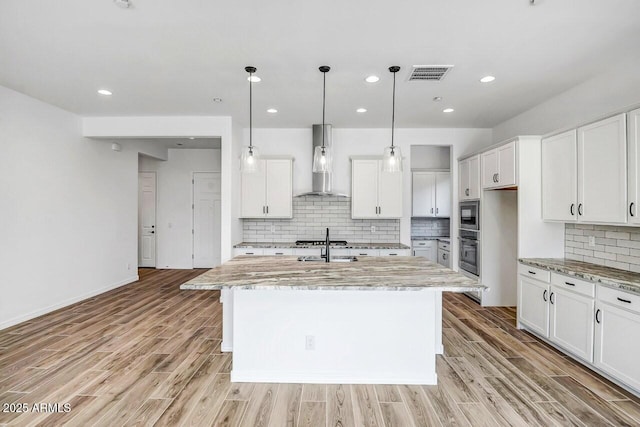 kitchen with light stone counters, wall chimney range hood, oven, and an island with sink