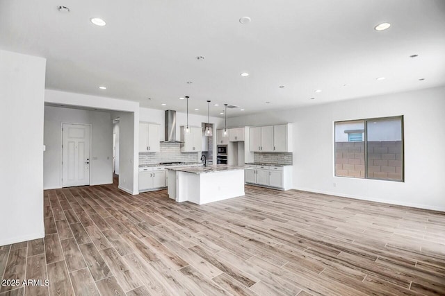 kitchen featuring pendant lighting, white cabinets, decorative backsplash, a center island with sink, and wall chimney exhaust hood