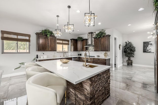 kitchen featuring dark brown cabinetry, wall chimney exhaust hood, sink, decorative light fixtures, and a center island with sink