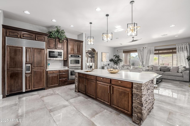 kitchen featuring dark brown cabinetry, sink, built in appliances, decorative light fixtures, and a kitchen island with sink