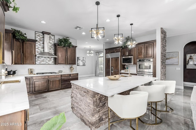 kitchen with dark brown cabinetry, built in appliances, a center island with sink, a notable chandelier, and wall chimney range hood