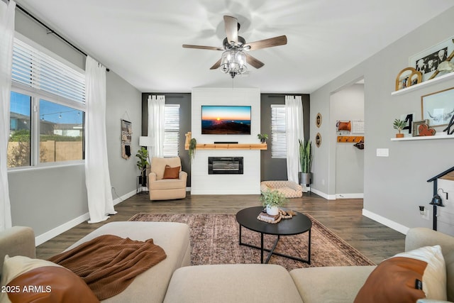 living area featuring a large fireplace, baseboards, dark wood-style floors, and ceiling fan