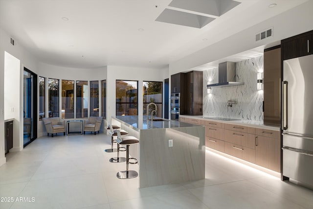 kitchen featuring wall chimney range hood, stainless steel appliances, tasteful backsplash, an island with sink, and a kitchen bar