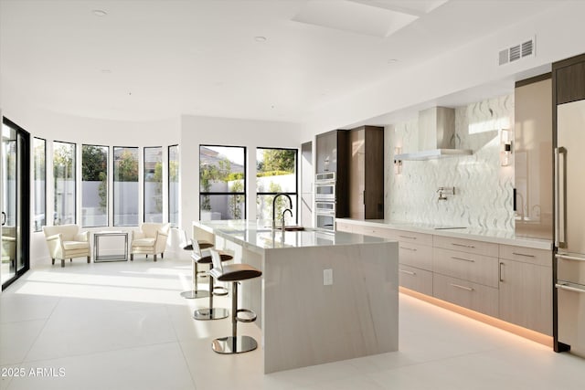 kitchen with wall chimney range hood, light tile patterned floors, a breakfast bar area, a kitchen island with sink, and stainless steel appliances