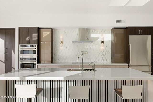 kitchen featuring wall chimney exhaust hood, a kitchen bar, dark brown cabinetry, sink, and double oven