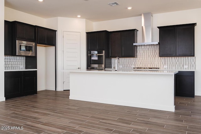 kitchen with backsplash, stainless steel appliances, wall chimney exhaust hood, and a center island with sink