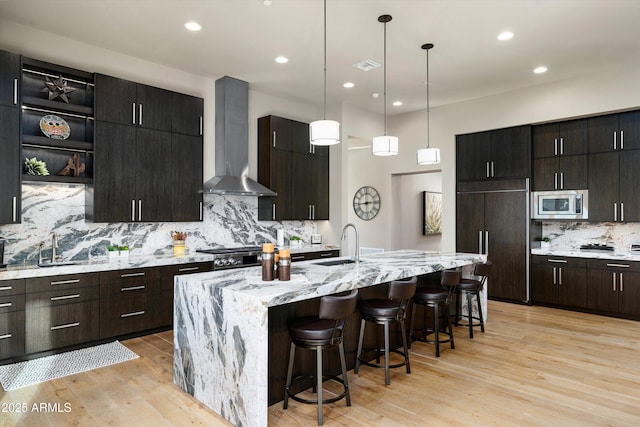 kitchen featuring a breakfast bar, tasteful backsplash, built in appliances, an island with sink, and wall chimney range hood