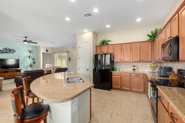 kitchen featuring light stone countertops, a kitchen island with sink, ceiling fan, sink, and black appliances